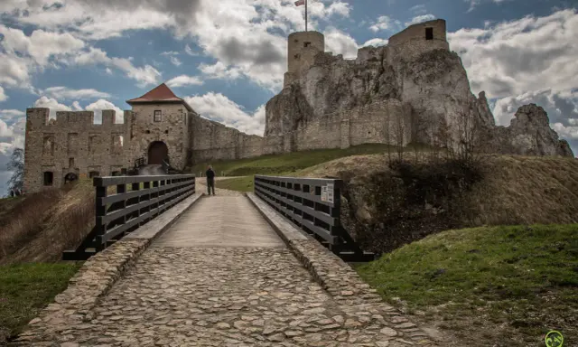 Kamienny most prowadzi do ruin zamku w Olkuszu. Na szczycie skalistego wzgórza widać wieżę z polską flagą.