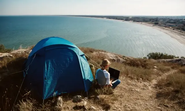 Niebieski namiot i osoba z laptopem na wzgórzu, z widokiem na lazurowe wybrzeże. Idealne miejsce na camping.