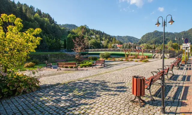 Słoneczna promenada Szczawnica z kamienną nawierzchnią, ławkami i latarniami, otoczona zielenią i górami.