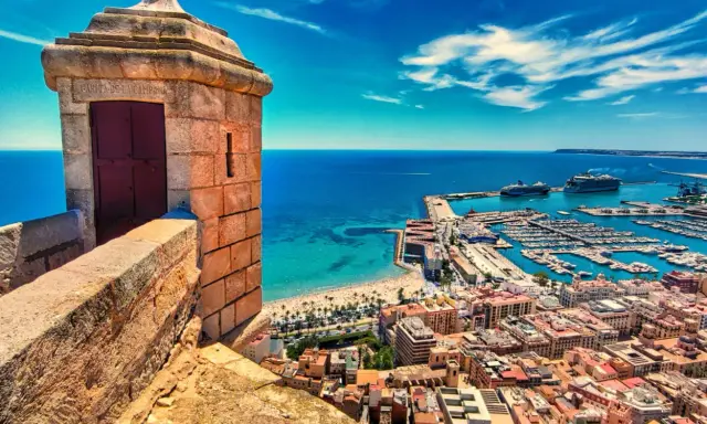 Alicante co zobaczyć: widok z zamku na port, plażę i miasto. Widać wieżyczkę strażniczą, jachty i statki wycieczkowe.