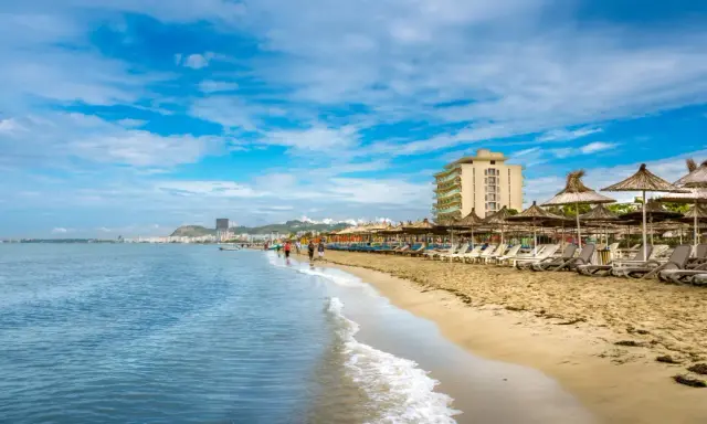 Słoneczna plaża w Durrës, Albania, z rzędami leżaków i parasoli. W tle widać hotel i miasto, a na pierwszym planie falujące morze. Idealne miejsce na wakacje i odkrywanie atrakcji.