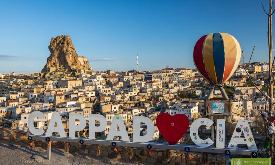 Cappadocia, Turcja zwiedzanie na własną rękę. Balon i napis "Cappadocia" na tle skalnych domów i twierdzy.
