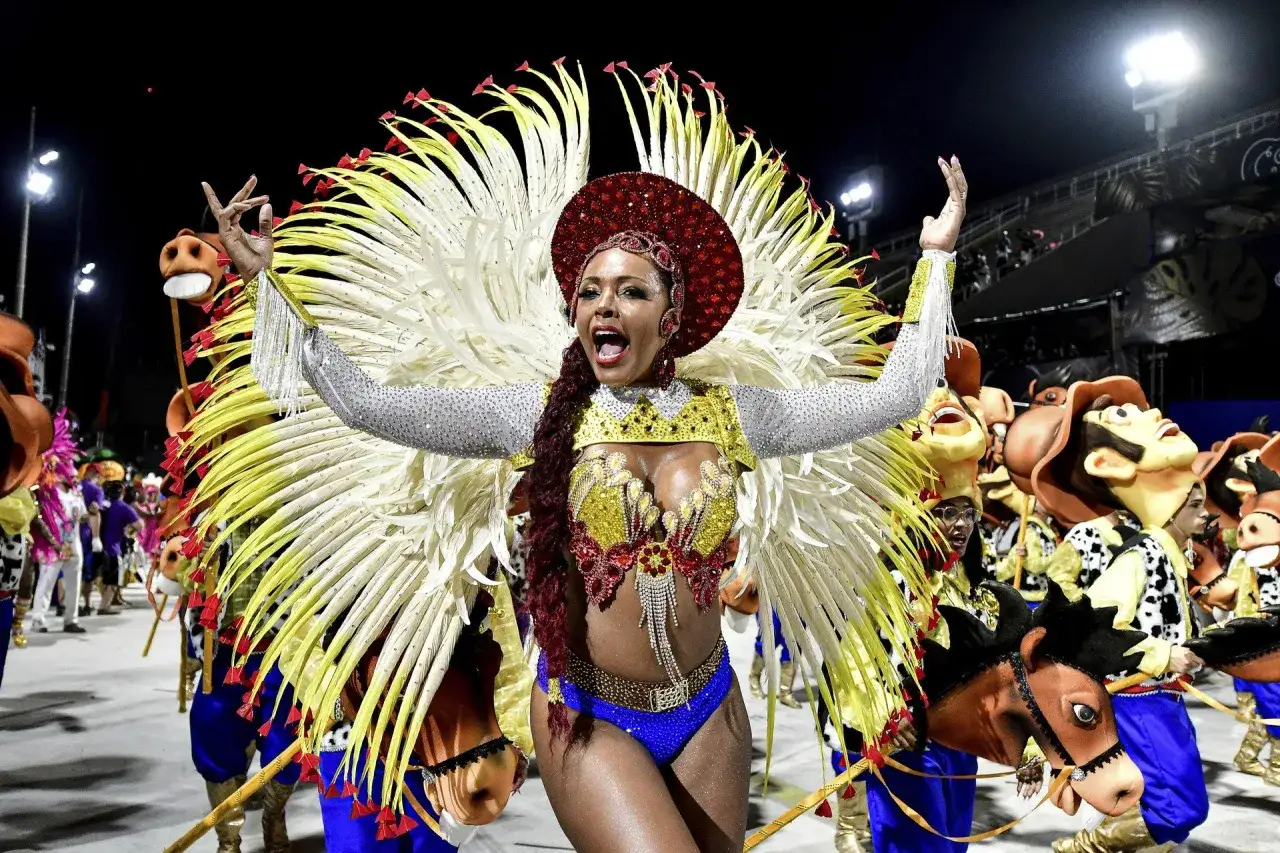 Tancerka w olśniewającym stroju z piór i cekinów na karnawale w Rio 2024. W tle barwne postacie i konie.