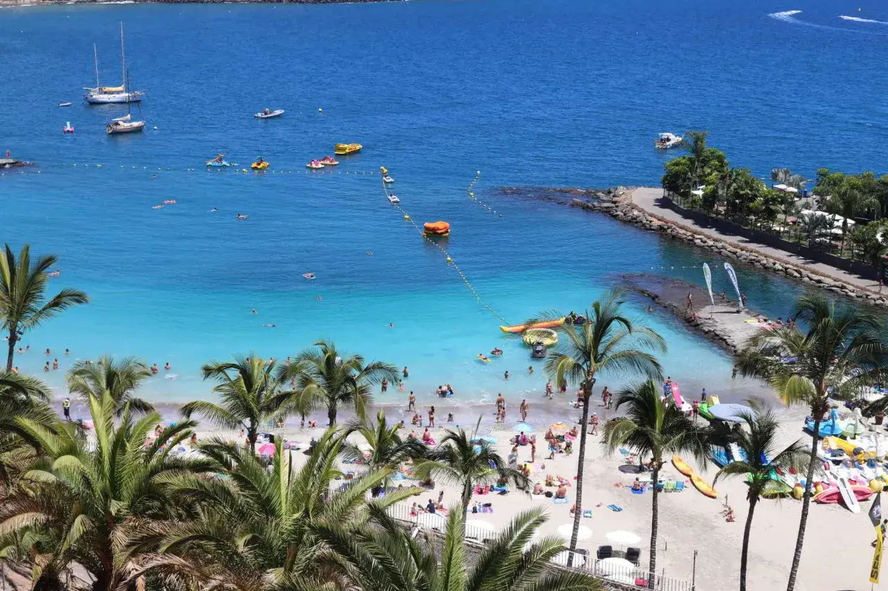 Słoneczne gran canaria plaże z palmami, turkusową wodą i ludźmi odpoczywającymi na piasku. Widać łodzie i sprzęt wodny.