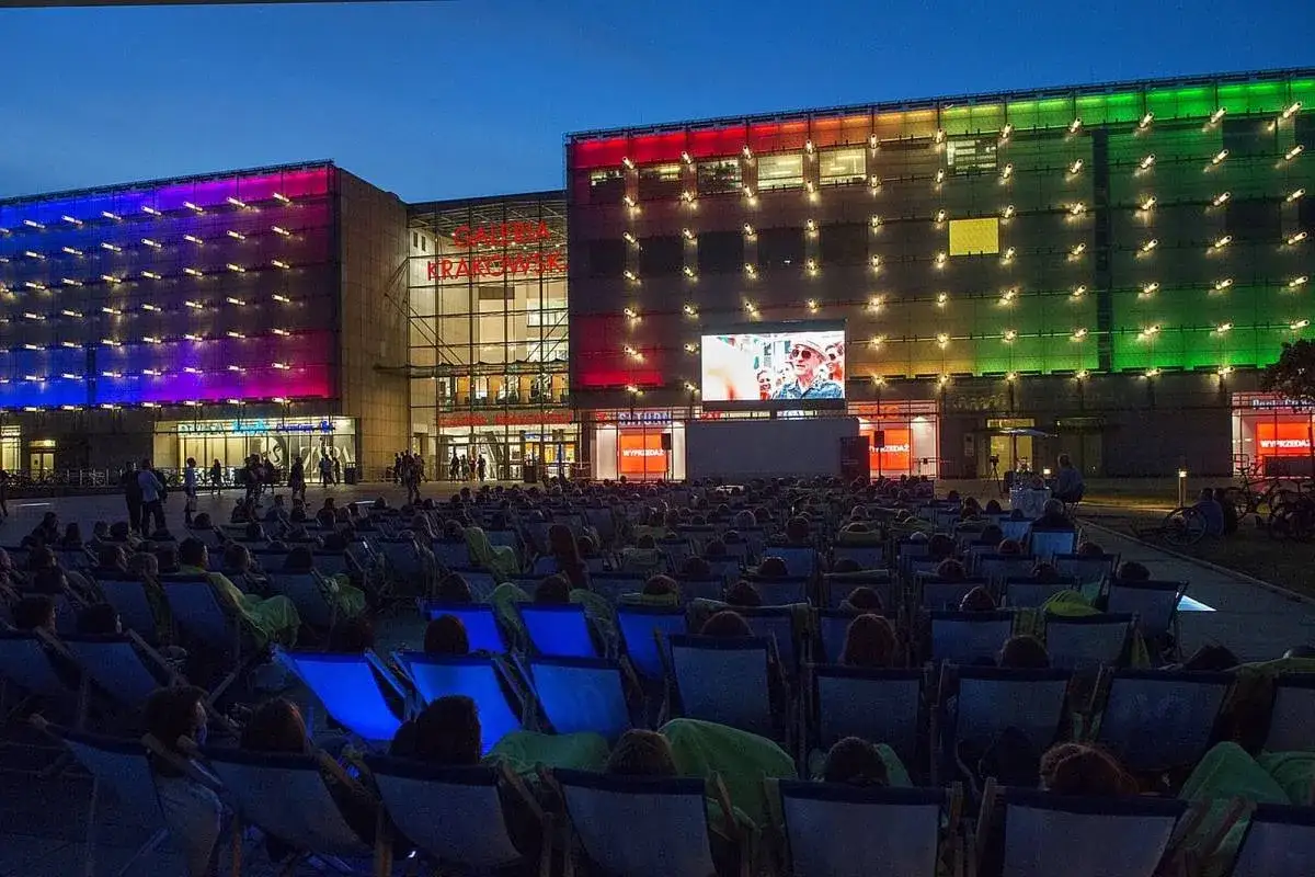 Wieczorne kino plenerowe przed Galerią Krakowską. Tłum widzów na leżakach ogląda film wyświetlany na wielkim ekranie. Budynek galerii rozświetlony kolorowymi światłami.