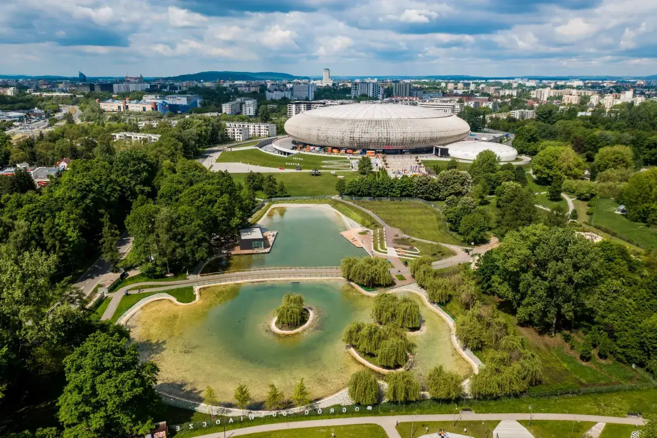 Malowniczy park lotników polskich z jeziorem i wyspą, w tle hala widowiskowa i miasto.