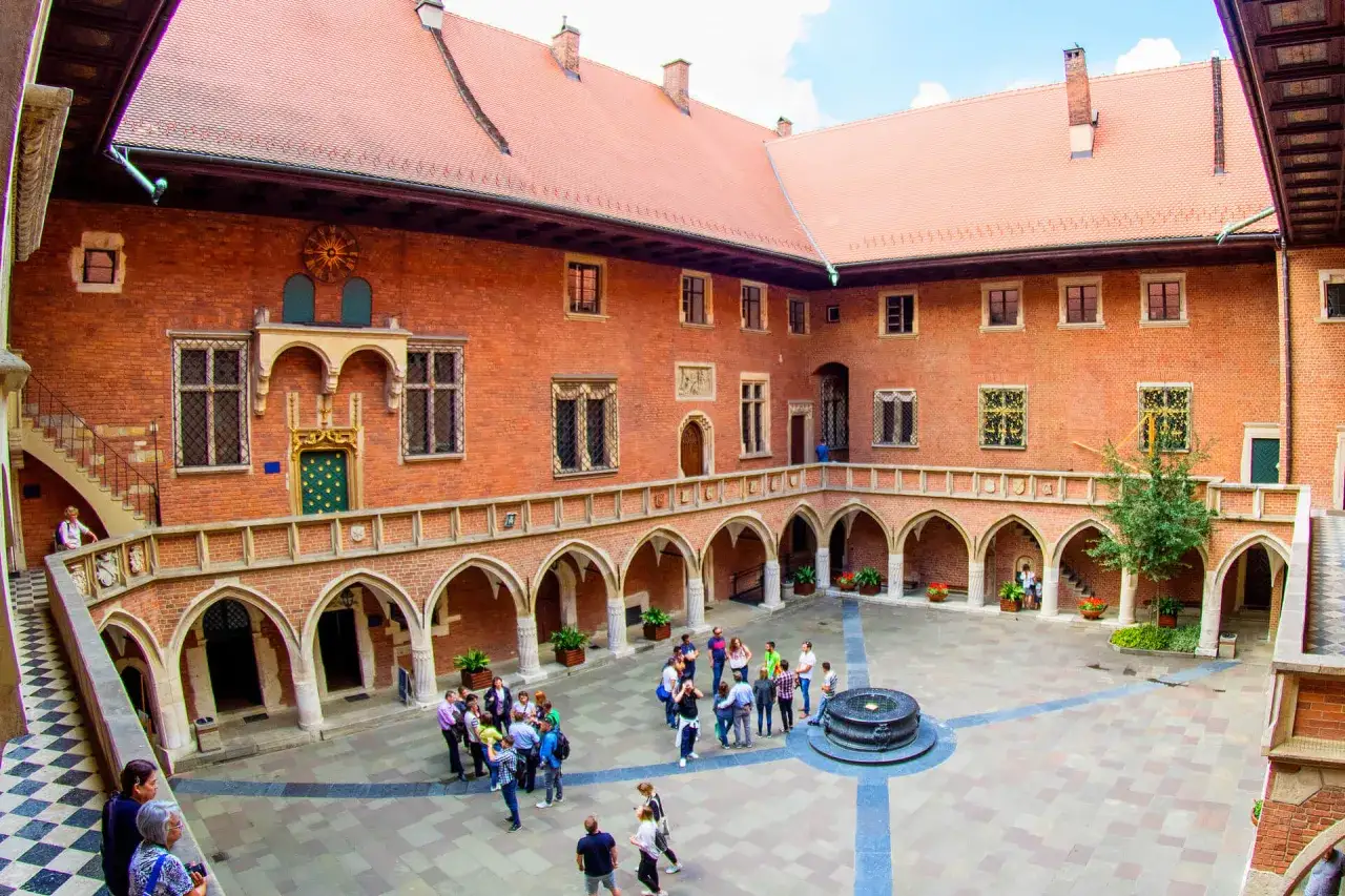 Dziedziniec Collegium Maius, z ceglanymi arkadami, schodami i grupami turystów zwiedzających historyczny budynek Uniwersytetu Jagiellońskiego.
