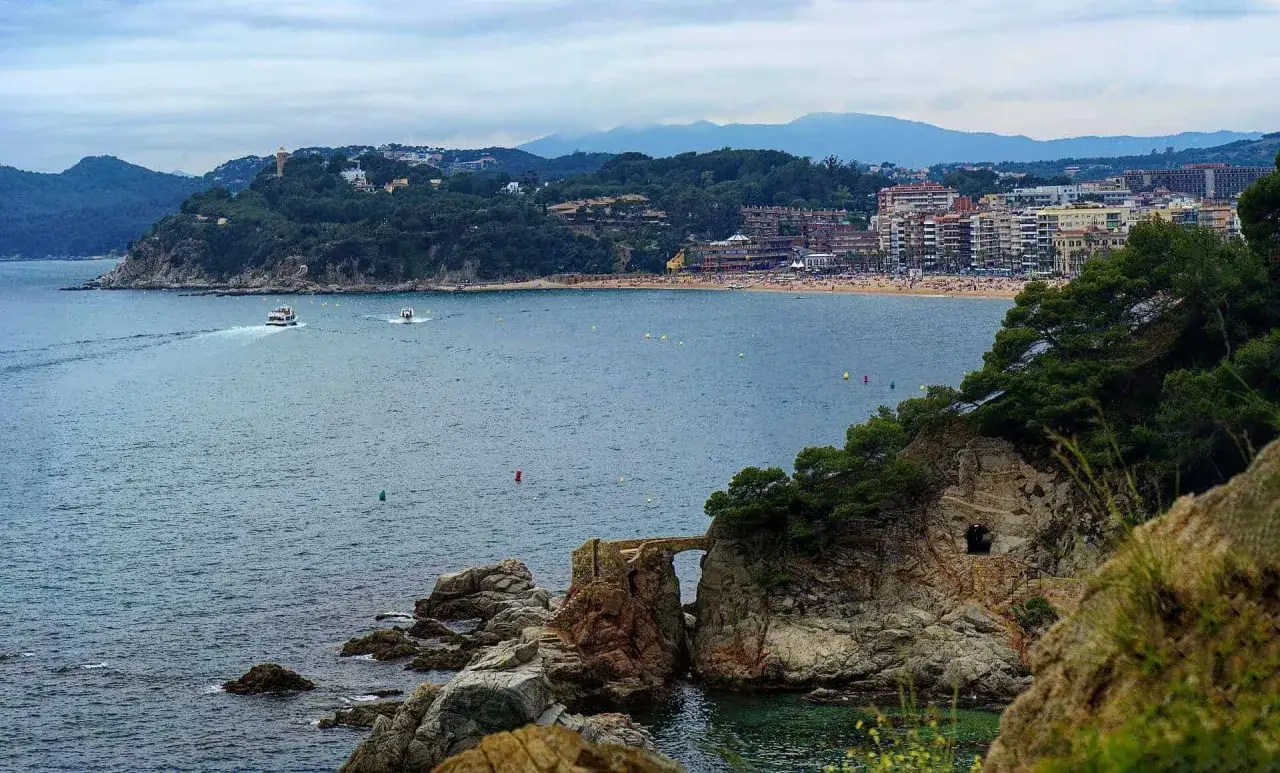 Widok na zatokę z łodziami, skalistymi wysepkami i mostkiem. W tle rozciągają się piękne costa brava plaże i zabudowania.