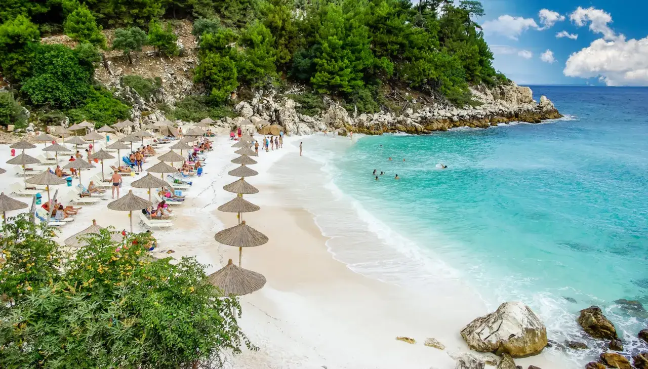 Piękna plaża z białym piaskiem i turkusową wodą. Rzędy parasoli i leżaków, ludzie odpoczywają i kąpią się. Thassos atrakcje w pełnej krasie.