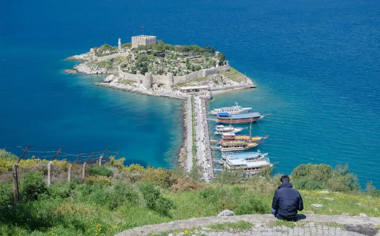 Widok na zamek na wyspie i przystań z łodziami w Kusadasi, Turcja. Mężczyzna siedzi na wzgórzu, podziwiając panoramę.