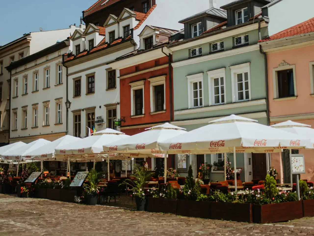 Kraków stare miasto restauracje z białymi parasolami Coca-Cola. Kolorowe kamienice tworzą tło dla letniego ogródka.