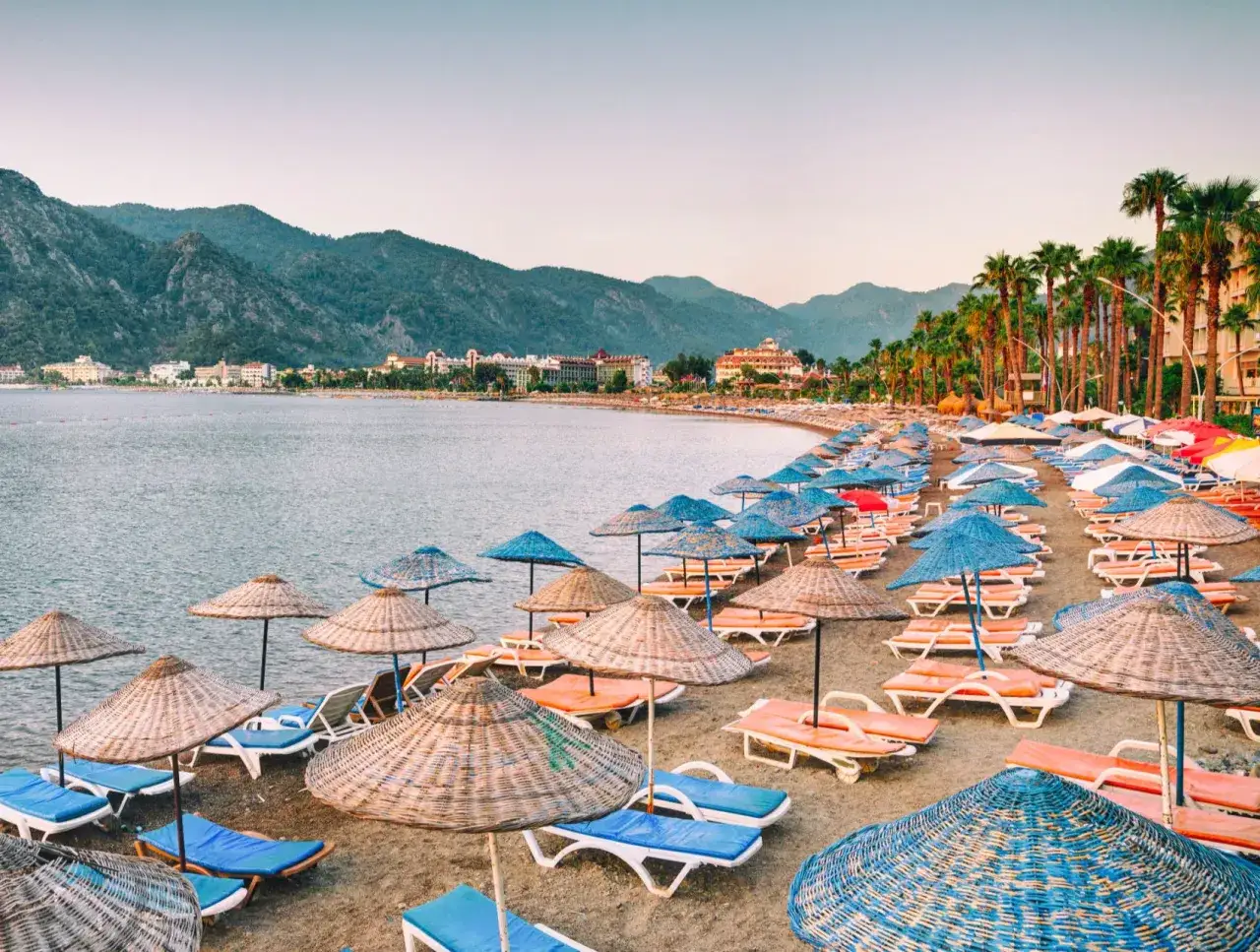 Plaża w Marmaris, Turcja, z rzędami leżaków i parasoli, w tle góry i hotele.