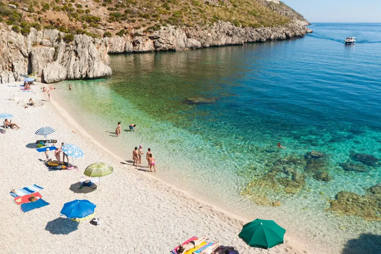 Słoneczna palermo plaża z turkusową wodą, kamienistym brzegiem i ludźmi odpoczywającymi pod parasolami.