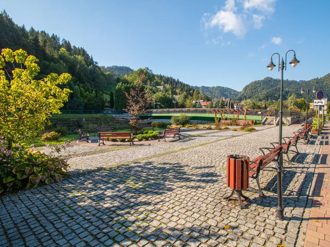 Słoneczna promenada Szczawnica z brukowanym chodnikiem, ławkami i latarniami, w tle góry i most.