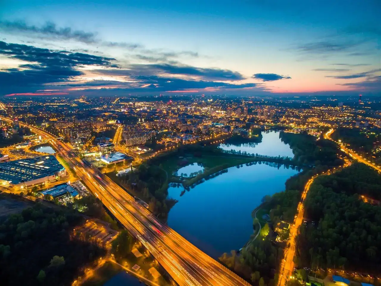 Zachód słońca nad miastem, gdzie autostrada przecina malowniczą dolinę trzech stawów w Katowicach, a światła miasta odbijają się w wodzie.