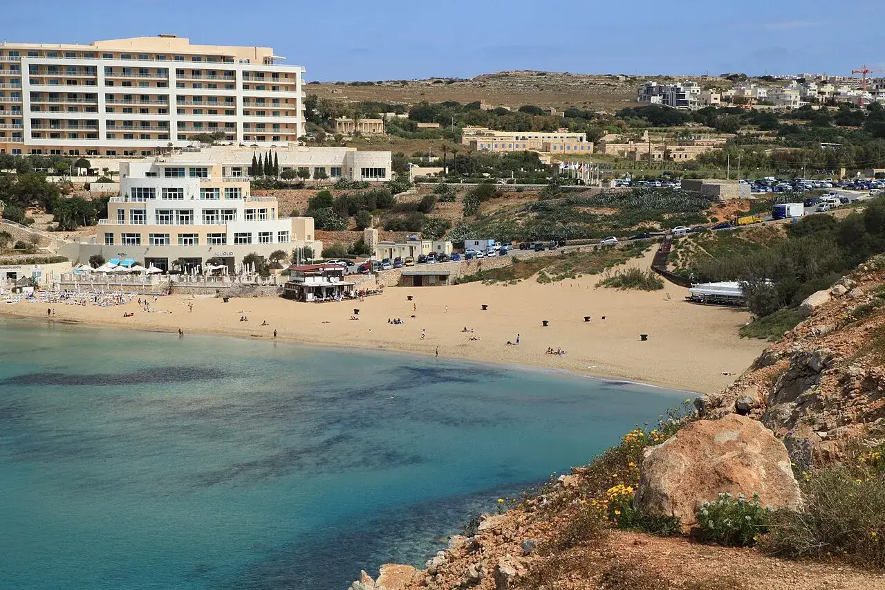 Malta plaże: piaszczysta zatoka z turkusową wodą, hotele i ludzie odpoczywający na piasku.