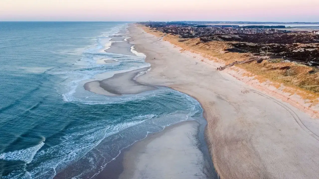 Szeroka, piaszczysta plaża na wyspie Sylt, z turkusowymi falami obmywającymi brzeg i wydmami porośniętymi trawą.