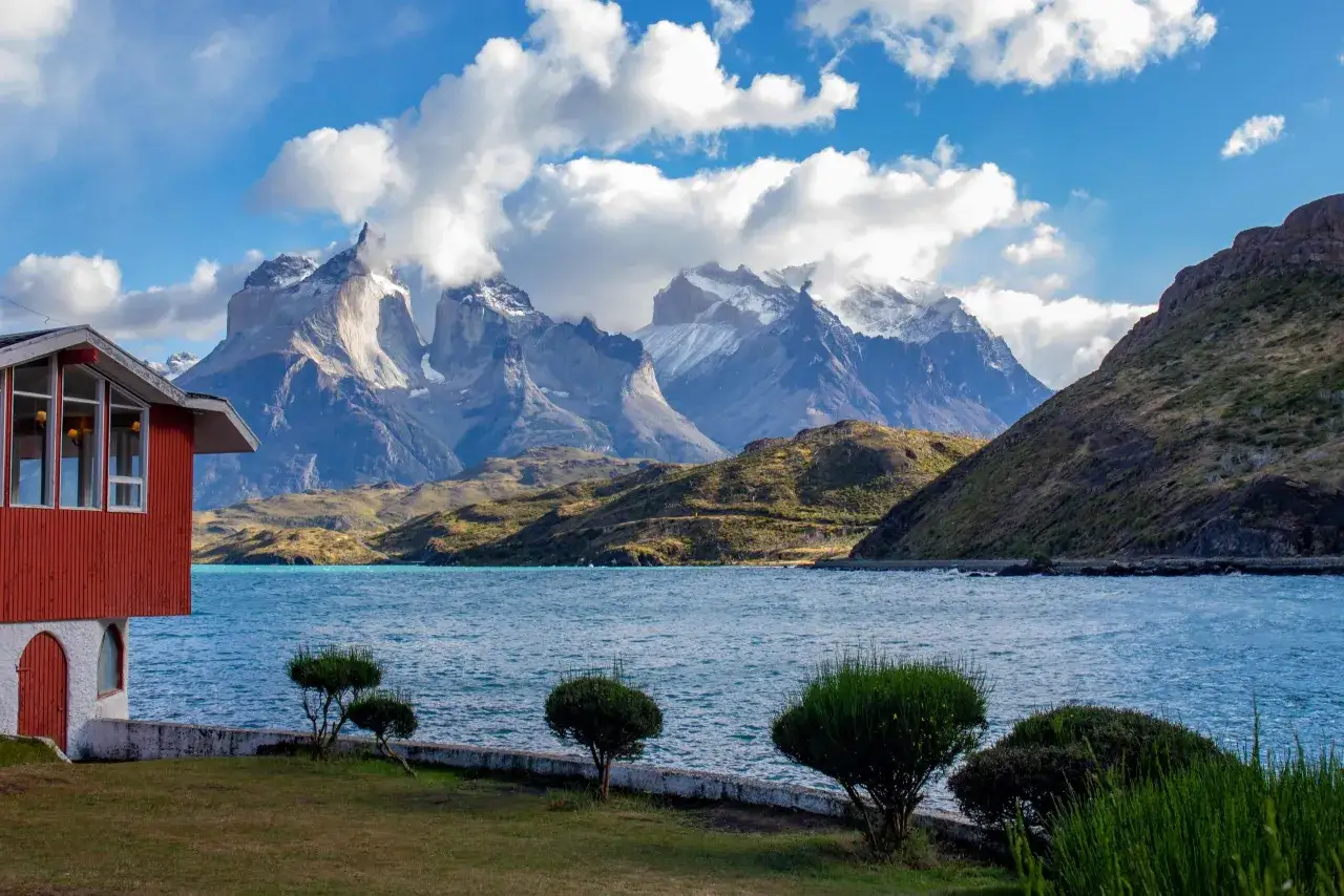 Widok na turkusowe jezioro i ośnieżone góry w Torres del Paine. Czerwony domek nad wodą to idealne miejsce, by zacząć odkrywać, co w Chile warto zobaczyć.
